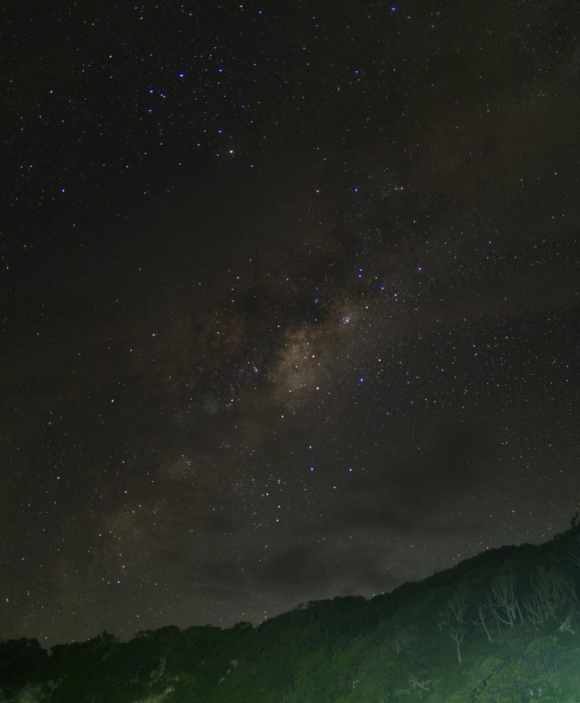 The Milky Way rising over Neds Beach on Lord Howe Island. The mountain range in the foreground has been lit up and is green below. Some wispy cloud cover can be seen approaching the Milky Way.