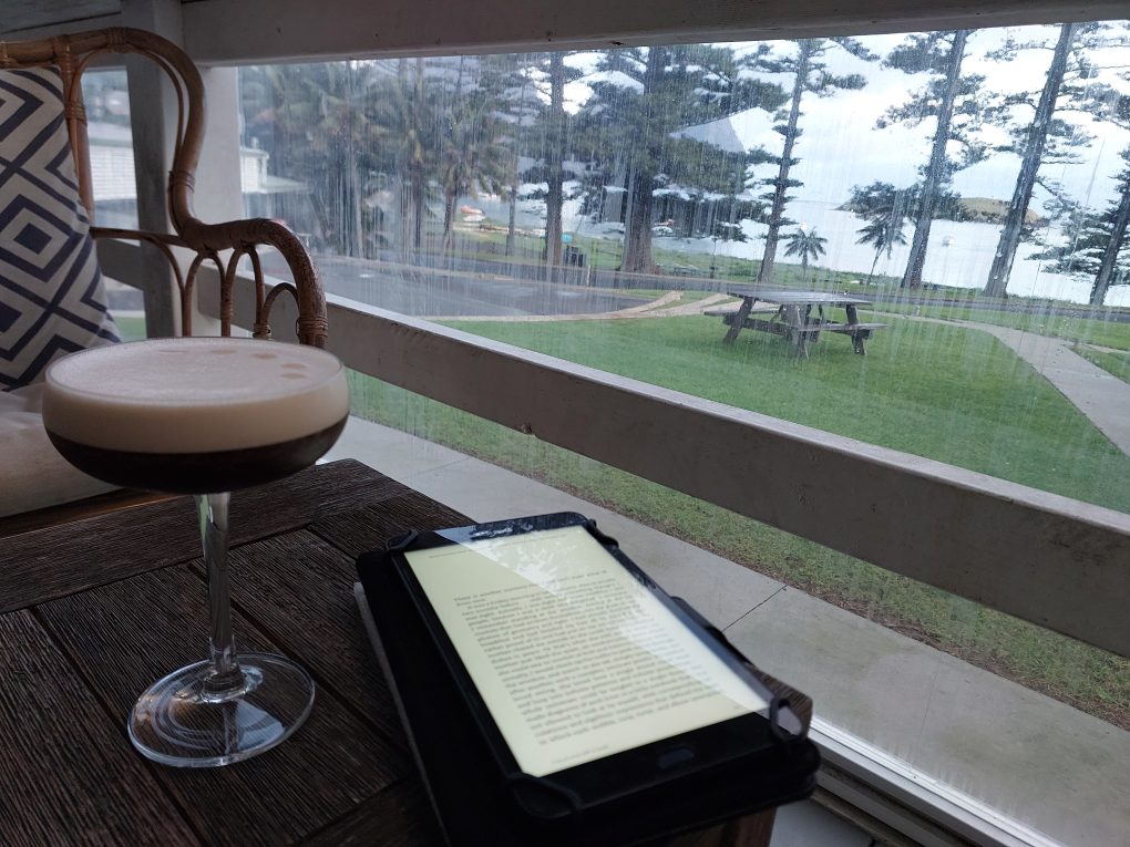 Pictured is a cocktail glass beside a tablet open to a book. Beyond can be seen the waters of the Lagoon
